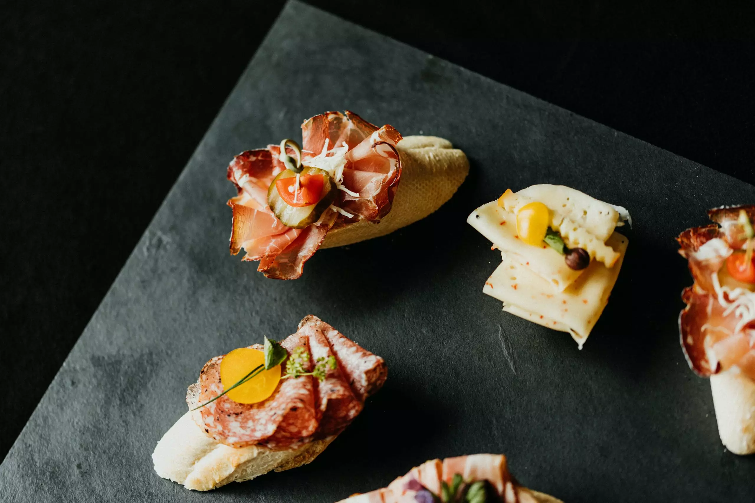 Verschiedene belegte Canapés mit Aufschnitt und Käse auf schwarzer Steinplatte, Cateringservice Präsentation