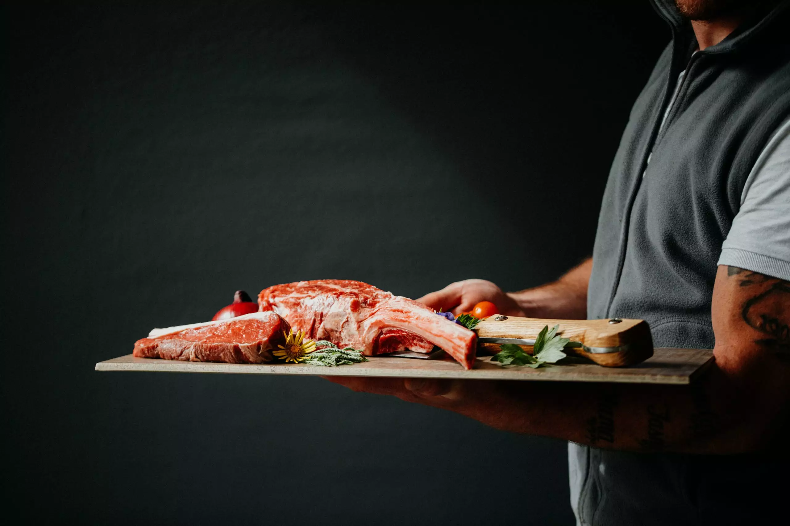 Mann hält Holzbrett mit rohem Steak, Kräutern und Tomate vor dunklem Hintergrund, Cateringservice Salzburg Oberösterreich