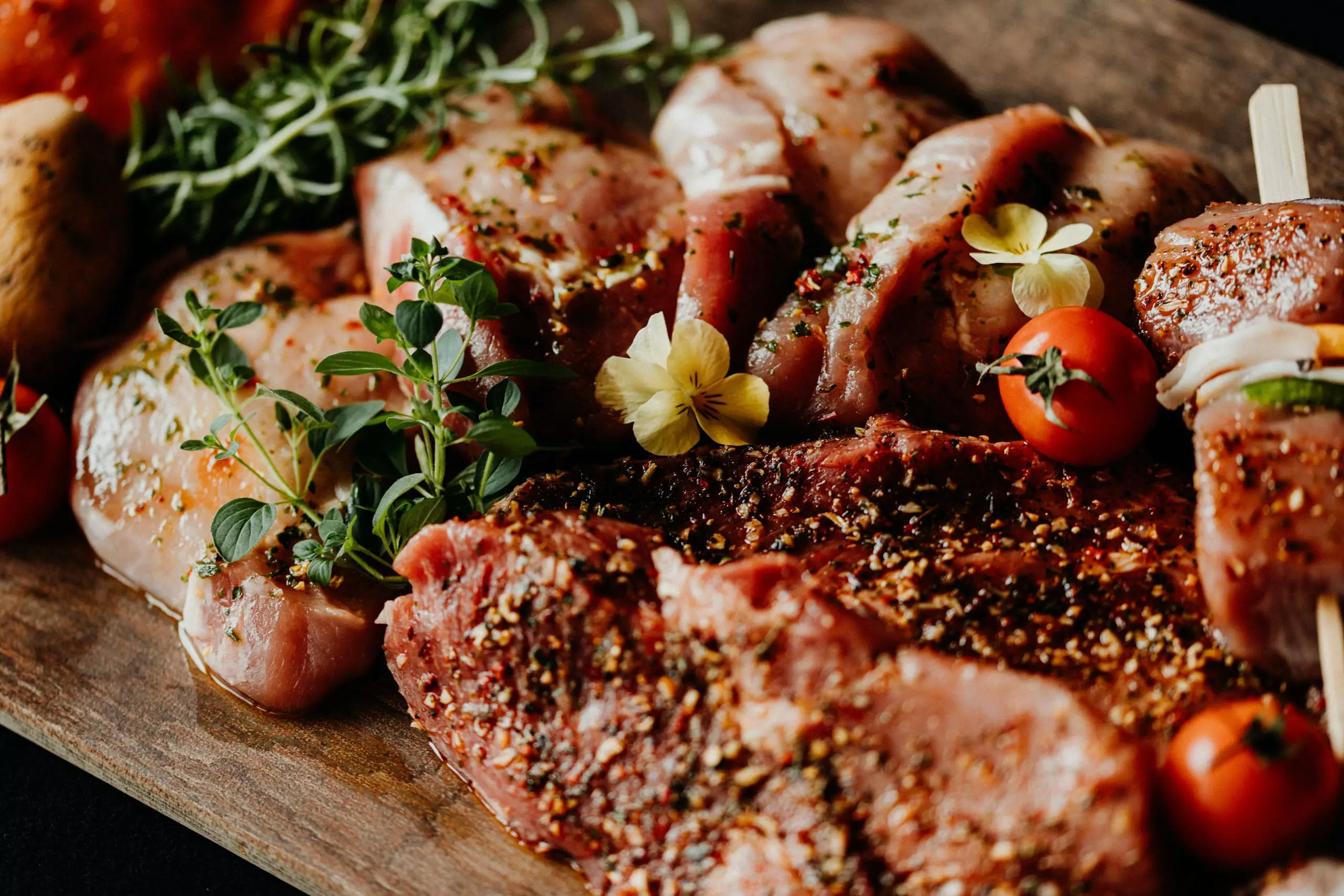 Würzig marinierte Fleischstücke mit frischen Kräutern, kleinen Tomaten und essbaren Blüten auf Holzbrett für Cateringservices