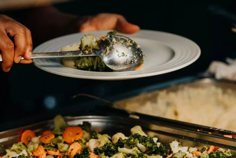 Hand schöpft gedünstetes Gemüse mit Brokkoli und Karotten auf weißen Teller in Betriebsrestaurant Cateringservice