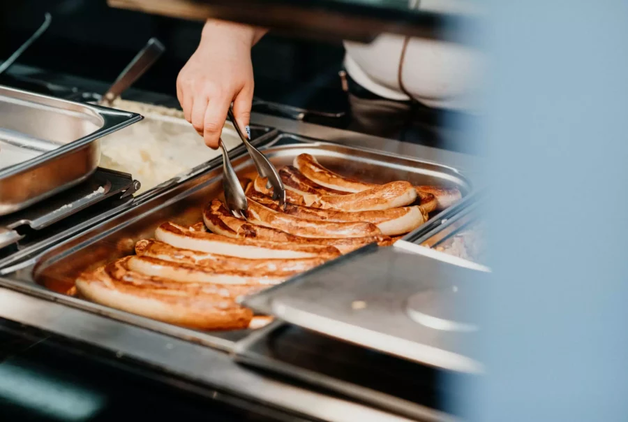Hand mit Zange nimmt Bratwürste aus einem Edelstahl-Buffetbehälter im Betriebsrestaurant Cateringservice