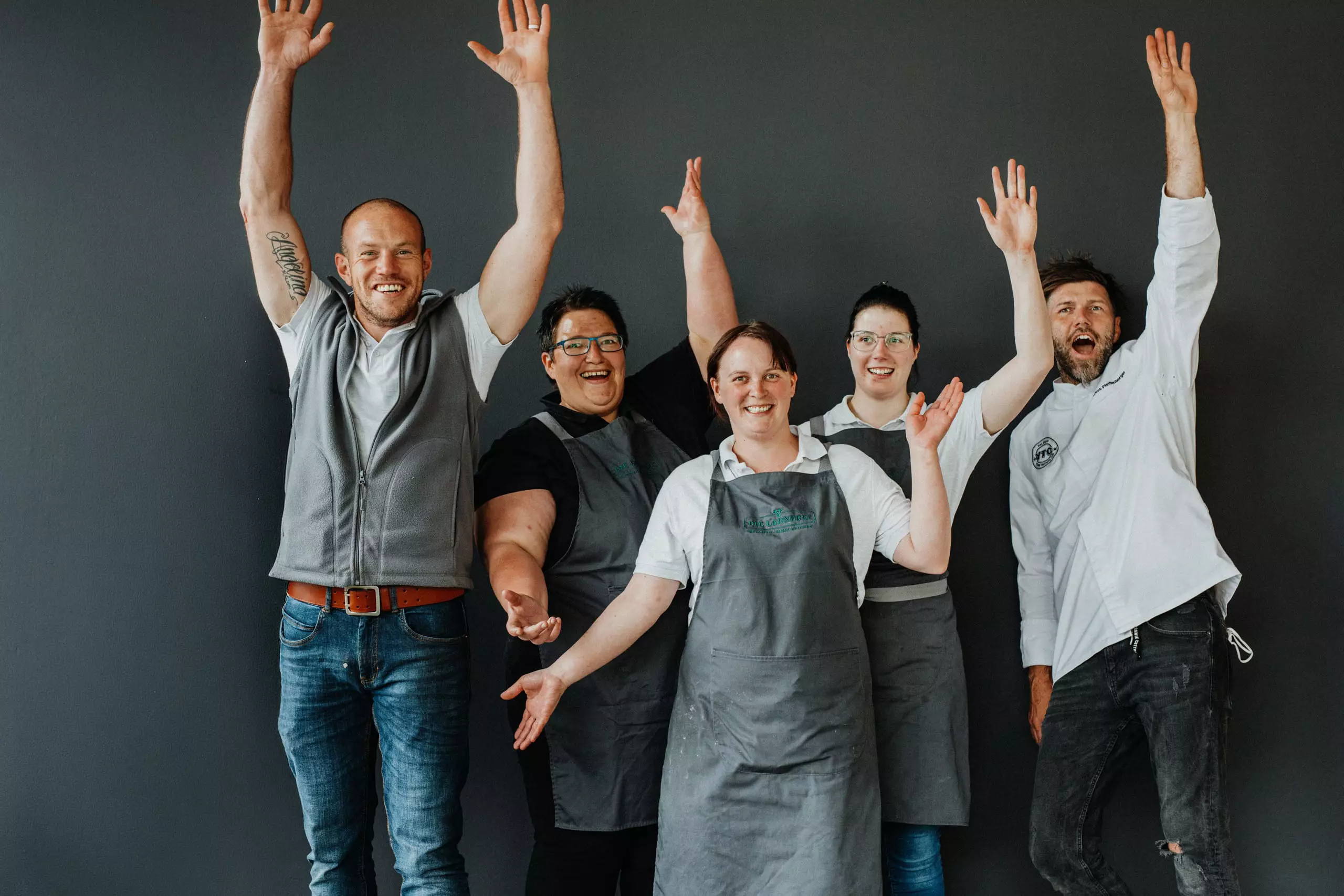 Fünf fröhliche Mitarbeiter eines Betriebsrestaurants in Arbeitskleidung vor grauer Wand mit erhobenen Armen und lachend.