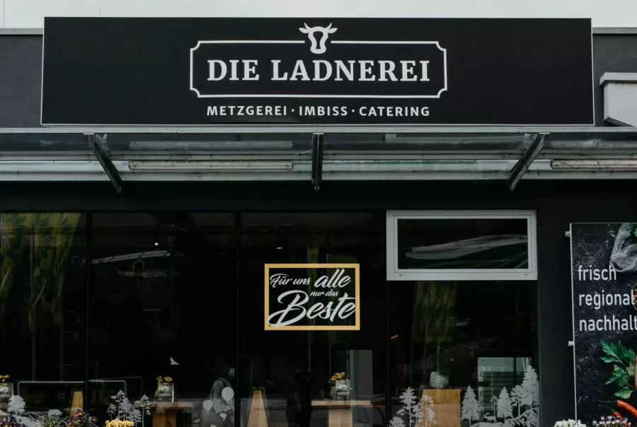 Eingang eines Betriebsrestaurants mit schwarzem Schild Die Ladnerei Metzgerei Imbiss Catering und dekorativen Pflanzkästen